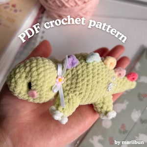May include: A light green crocheted dinosaur with colorful accents, including pink, purple, and yellow. The dinosaur has a white ribbon with a small flower and the text "PDF crochet pattern".