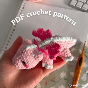 May include: A handmade pink crochet dragon with bright pink wings and light green accents. The dragon is held in a hand. The text "PDF crochet pattern" is displayed above the dragon.