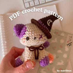 Puede incluir: Un oso de ganchillo hecho a mano con un sombrero de bruja. El oso es de color crema con detalles morados y naranjas, detalles cosidos y un lazo marrón. El sombrero es marrón con un borde morado y una luna creciente. El texto en la imagen dice "PDF crochet pattern".