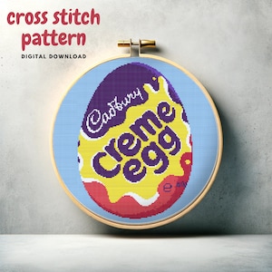 May include: A cross stitch pattern of a Cadbury Creme Egg. The egg is purple and yellow with the words "Cadbury Creme Egg" in purple letters. The pattern is in a wooden embroidery hoop.