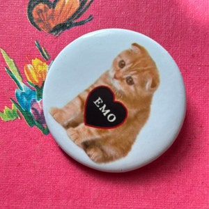 May include: A white button with a picture of a ginger kitten holding a black heart with the word "EMO" written on it.