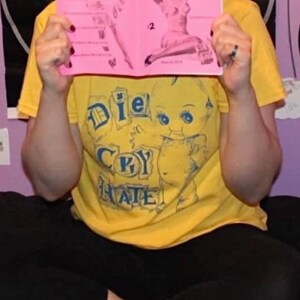 May include: A person wearing a yellow t-shirt with a blue graphic of a baby doll holding a heart and the text "Die Cry Hate". The person is holding a pink booklet with the text "The Escapist Artist" and the number "53" on the cover.