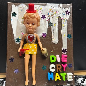 May include: A mixed media artwork featuring a doll with a red top hat, holding a toy gun and a skull. The doll wears a yellow skirt with white hearts. The words "DIE CRY HATE" are spelled out with colorful letter beads, with flower and sprinkle embellishments.
