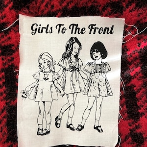 May include: A white fabric patch with black text that reads "Girls To The Front". The patch features a black and white illustration of three girls wearing dresses.