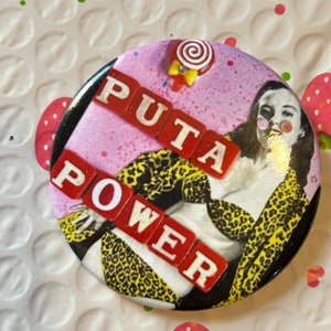May include: A round button with a pink background and a woman wearing a leopard print outfit. The words "PUTA POWER" are spelled out in red letters on the button.