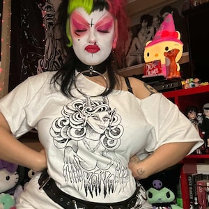 May include: White t-shirt with a black and white graphic of a horned woman with the text "Katya". The shirt is paired with a black belt with silver rings. The person is wearing a black choker and a tiara.