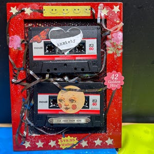 May include: A red rectangular frame containing two black Maxell UR 60 cassette tapes decorated with stickers including a heart labeled Anxiety, a butterfly, a moon face, and text reading 42 Golden Hits and Far Away From Here, with loose tape.