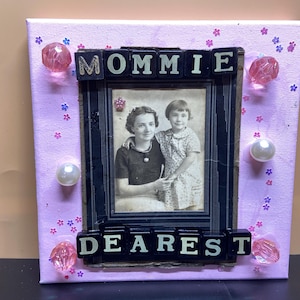 May include: A framed photograph with the words "MOMMIE DEAREST" in black letter tiles. The frame is on a pink canvas decorated with small flower and gem embellishments. The photograph is of two people.