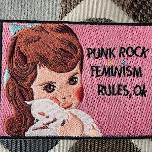 Puede incluir: Parche bordado con fondo rosa y borde negro. El parche presenta una ilustración de estilo vintage de una niña con cabello castaño sosteniendo un gato blanco. El texto dice "PUNK ROCK FEMINISM RULES, OK."