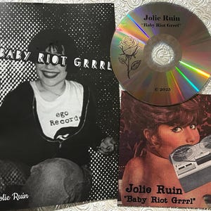 May include: A promotional image featuring a CD, a poster, and a card. The CD and card display the text "Jolie Ruin" and "Baby Riot Grrrl". The poster features a black and white photo of a person wearing a shirt that says "ego Record".