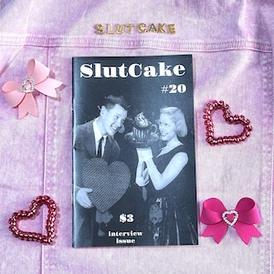 May include: A black and white magazine cover with the title "SlutCake" and the number "#20" printed in white. The cover features a photo of a couple in a vintage setting, holding a heart-shaped box. The text "$3 interview issue" is printed at the bottom of the cover.