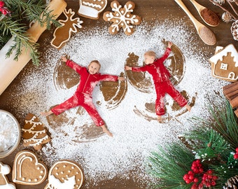 May include: Two children in red pajamas make snow angels in a white powder on a wooden surface.  Gingerbread cookies in the shape of a house, a snowflake, a reindeer, a Christmas tree, and a stocking are scattered around the children.