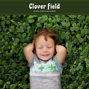 May include: A young boy with brown hair and a grey shirt with a blue and white graphic lies on his back in a field of green clover.