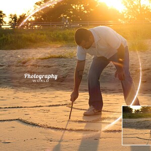 May include: A person draws a heart shape in the sand on a beach at sunset. The sun is shining brightly and creating a lens flare effect. The person is wearing a white shirt and blue jeans.
