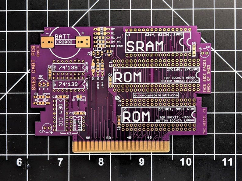 Puede incluir: Una placa de circuito impreso (PCB) de color p&uacute;rpura para un cartucho de juego de Super Nintendo Entertainment System (SNES). La placa tiene un soporte para bater&iacute;a, un chip 74'139, un chip ROM de m&aacute;scara 27C801, un chip ROM 27C160 y un chip SRAM 6261. La placa est&aacute; etiquetada como "SNES CART PCB" y "Mousebite Labs".