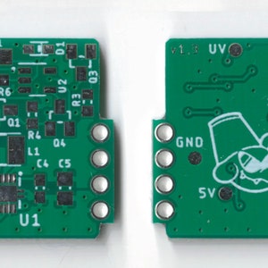 May include: A green circuit board with electronic components, including resistors, capacitors, and an integrated circuit. The board has a white outline of a cartoon mouse wearing a hat and the text "v1.3 UV" and "EN".