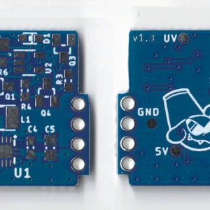 May include: A blue circuit board with various components, including resistors, capacitors, and an integrated circuit labeled "U1". The board has a white outline and a white text label "v1.3" in the upper right corner. The board also features a white cartoon illustration of a creature with a hat.
