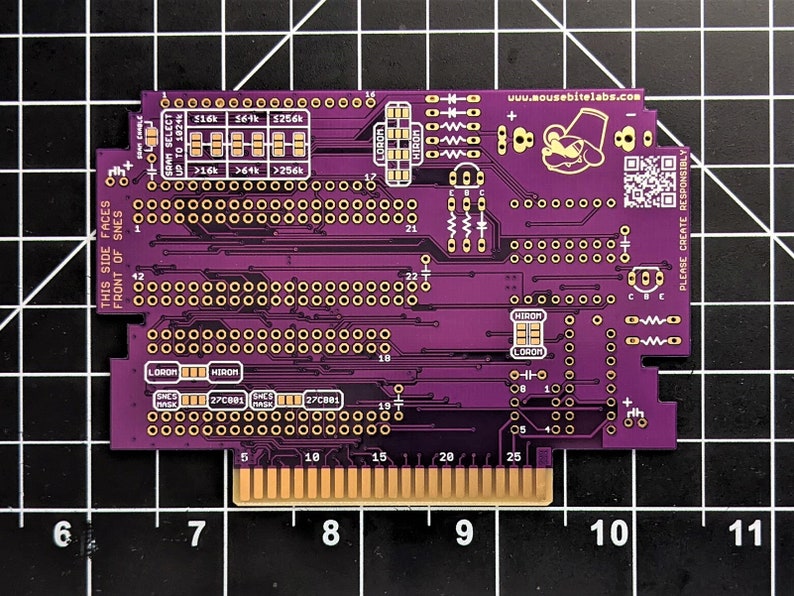 Puede incluir: Una placa de circuito impreso de color p&uacute;rpura con componentes y texto de color dorado. La placa tiene una variedad de conectores y chips. El texto en la placa incluye "THIS SIDE FACES FRONT OF SNES", "LOROM", "HIROM", "SNES", "27C001" y "27C801". La placa tambi&eacute;n tiene un peque&ntilde;o logotipo con un personaje de dibujos animados que lleva un sombrero y el texto "www.mousebitelabs.com".