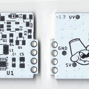 May include: A white circuit board with black components and text, including "v1.3", "UV", "EN", "SW", "GND", "VCC", "5V", and "U1". A cartoon dog with a hat is printed on the board.