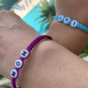 May include: Two friendship bracelets with the names "MOM" and "ARI" spelled out in white beads on purple and blue string.