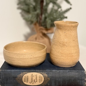 May include: A set of two handmade ceramic pieces, a small bowl and a vase, both in a speckled beige colour. The pieces are sitting on top of two vintage books.