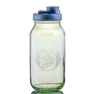 May include: A clear glass water bottle with a blue lid. The bottle has a vintage design with the words "BRIT & AZURE" and a mountain and wheat design. The bottle is approximately 7 inches tall.
