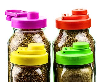 May include: Four glass jars filled with seeds, each topped with a brightly colored lid. The lids are pink, red, yellow, and green. The jars are filled with different types of seeds, and the background is white.
