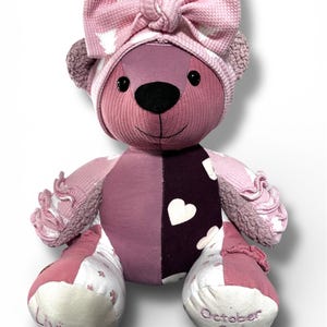 May include: A plush teddy bear crafted from various pink and dark purple fabrics with heart patterns and textured sections. The bear wears a matching pink knotted bow headband. The left foot is embroidered with Liviana and the right with October 2nd 2024.