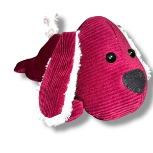 May include: A soft toy dog made from deep red corduroy fabric. The dog features black button eyes, a black nose, and white fluffy trim on the ears and tail. A charming children's toy.