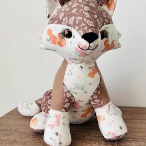 Könnte beinhalten: Ein Stofftier-Fuchs mit einem braunen, floral gemusterten Kopf und Ohren sowie einem weißen Gesicht und Pfoten mit Eichhörnchen- und Hasenprints. Der Fuchs hat große Augen und einen lächelnden Ausdruck.