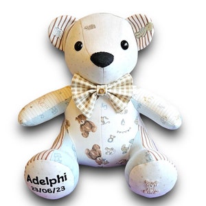 May include: A white patchwork teddy bear with brown and white striped ears. The bear is wearing a brown and white checked bow tie. The bear has a black nose and black eyes. The bear's paws are embroidered with a brown bear design. The bear's foot is embroidered with the name "Adelphi" and the date "23/06/23".
