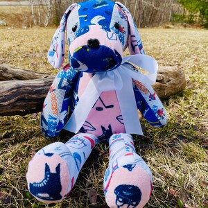 Peut inclure: Un jouet en peluche pour chien fait main à partir de tissus recyclés. Le chien est assis sur un lit d'herbe verte. Le chien porte un ruban blanc autour du cou. Le corps du chien est rose et ses oreilles et ses pattes sont bleues et blanches.