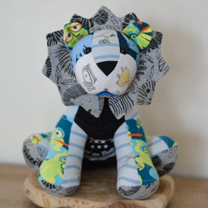 May include: A handmade stuffed lion toy made from recycled fabric. The lion has a blue and grey striped body, a black face, and a grey mane with black leaf patterns. The lion is sitting on a wooden surface.
