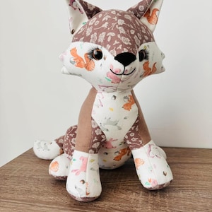 Könnte beinhalten: Ein braunes und weißes Plüschfuchs-Spielzeug mit einem Waldtier-Druck. Der Fuchs sitzt mit gekreuzten Beinen und hat einen freundlichen Ausdruck.
