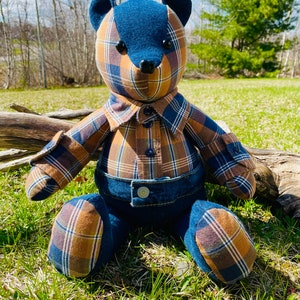May include: A handmade plush teddy bear wearing a plaid shirt and denim dungarees. The bear is sitting on a bed of green grass.
