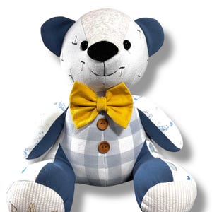 Op de afbeelding: Een handgemaakte teddybeer met een wit en blauw geruit lichaam, marineblauwe oren en een gele vlinderdas. De beer heeft een zwarte neus en knoopogen. De poten van de beer zijn geborduurd met de letters "MAC".