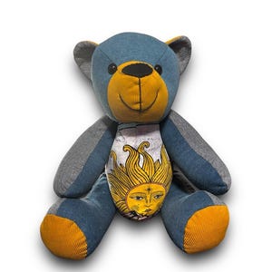 May include: A handmade teddy bear with a blue body, grey arms and ears, and yellow accents on the snout and feet. The bear's belly features a sun design with a face. The bear is made of soft fabric and has a smiling face.