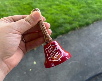 May include: A hand bell with a brown, wood-grain textured handle and a glossy red metal bell. The text THE SALVATION ARMY is printed in white on the side of the bell, accompanied by a small registered trademark symbol near the bottom edge.