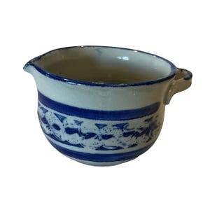 May include: A white ceramic pitcher with a blue rim and a blue band with a floral design. The pitcher has a small handle and a spout for pouring. The pitcher is likely used for serving liquids.