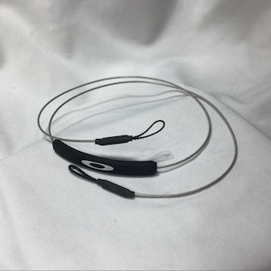May include: Black and grey eyewear retainer with the Oakley logo. The retainer features a black rubber piece with the Oakley logo and grey wire cords with loops on each end. The retainer is designed to secure eyewear.