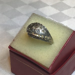 May include: A silver ring with a delicate, intricate design featuring small, clear stones. The ring is displayed in a red velvet box.