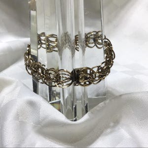 May include: A gold-toned metal bracelet featuring a multi-layered chain design with interconnected circular links. The bracelet includes a rectangular box clasp mechanism with a small round button release, creating a continuous circular structure.