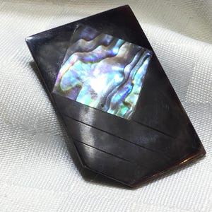 Peut inclure: Une broche rectangulaire présentant une surface polie brun foncé avec une incrustation centrale en forme de losange en nacre d'ormeau irisée. La partie inférieure de la broche affiche trois lignes diagonales gravées parallèlement.