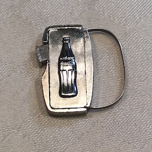 May include: A silver bottle opener featuring a Coca-Cola bottle design. The opener has a ring handle and a bottle opener blade.