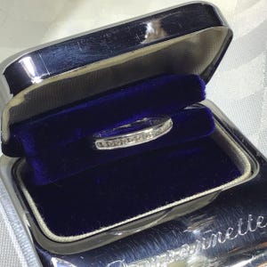 Peut inclure: Une bague anneau argentée avec une rangée de petites pierres transparentes serties en rail, présentée dans un écrin à bijoux doublé de velours bleu foncé avec un couvercle métallique portant le mot "pernette" visible en écriture cursive.