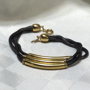 Peut inclure: Un bracelet or et noir avec un design à trois brins. Le bracelet présente des accents cylindriques dorés et des fermoirs dorés. Les brins sont noirs et ont l'apparence d'un serpent. Le bracelet est présenté sur une surface blanche.