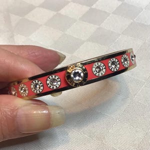 Peut inclure: Un bracelet jonc en or avec une base en émail rouge corail et des accents en émail blanc. Le bracelet est décoré de petits strass et présente un motif répétitif de chiffres.