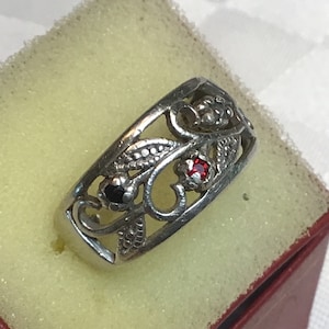 May include: A silver ring with a delicate filigree design featuring a red and black gemstone. The ring is set on a red velvet background.