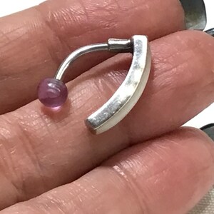 May include: A curved belly button ring with a silver-coloured metal bar and a small, round, purple gemstone. The ring has a curved design with a flat, rectangular end.