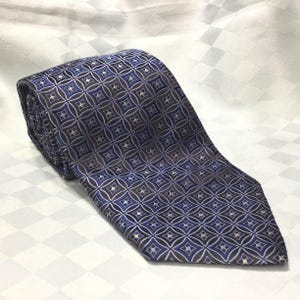 May include: Rolled-up tie with a geometric pattern in shades of blue, grey, and silver. The repeating design is made up of square and diamond shapes. The tie is made of a smooth fabric, suitable for formal wear.